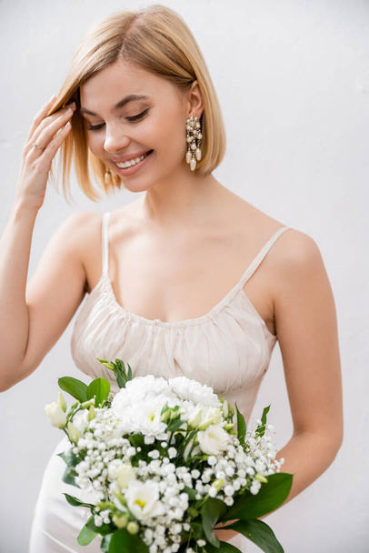 vrolijke en blonde bruid in trouwjurk met boeket op grijze achtergrond, witte bloemen, bruids accessoires, geluk, speciale gelegenheid, mooi, vrouwelijk, gelukzalig  - Foto, afbeelding