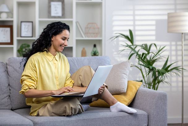 Młoda kobieta pracująca z laptopem siedzącym na kanapie w domu, odnosząca sukcesy kobieta z Ameryki Łacińskiej w salonie pisząca na klawiaturze komputera pracująca zdalnie online. - Zdjęcie, obraz