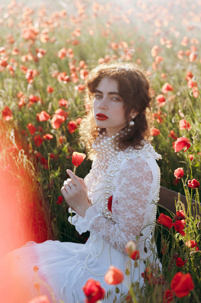 A beautiful girl in a vintage white dress, as a model, at a fashion photo shoot in a poppy field at sunset. - Foto, imagen