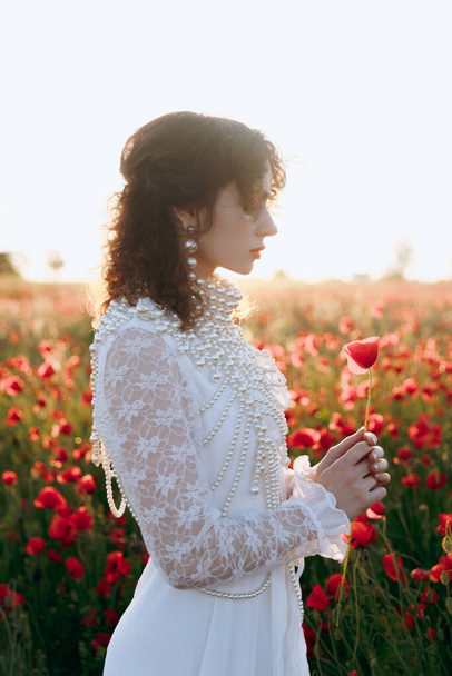 A beautiful girl in a vintage white dress, as a model, at a fashion photo shoot in a poppy field at sunset. - Foto, Bild
