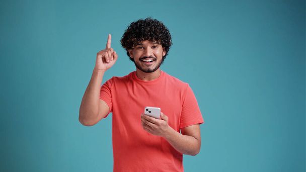 Young Hispanic or Arab man using smartphone standing very happy pointing with hand and finger showing up, in coral t-shirt on blue studio background. - Foto, imagen