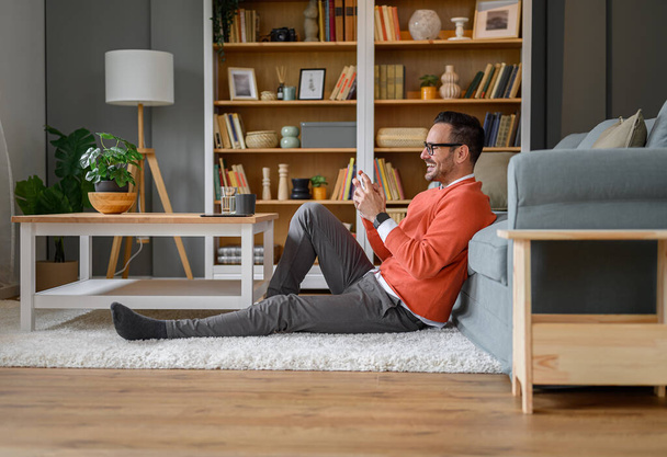 Vista lateral del joven alegre jugando videojuego por teléfono inteligente mientras está sentado en la alfombra en casa - Foto, imagen
