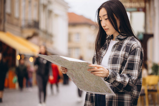 旧市街の通りに地図を持っている美しいブルネットの韓国人女性の肖像画。ヨーロッパを旅行するアジアの女性観光客やビジネス女性. - 写真・画像
