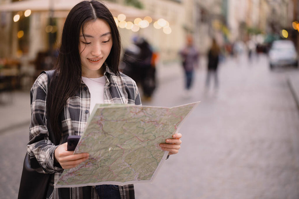 Portrait of a beautiful brunette Korean woman holding a map on the streets of the old city. Asian woman tourist or business lady traveling in Europe. - Photo, Image