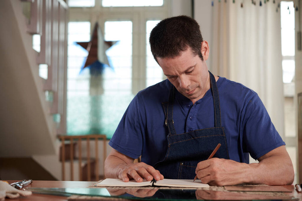 Glaser konzentriert sich vor der Fertigung auf das Zeichnen von Skizzen, während er in seiner Werkstatt am Tisch sitzt - Foto, Bild