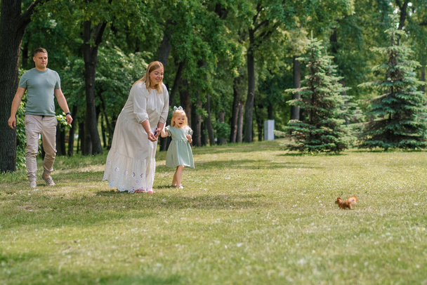 Ευτυχισμένη οικογένεια σε μια βόλτα στο πάρκο, η μητέρα δείχνει στην κόρη της ένα σκίουρο να πηδάει στο γρασίδι. Το παιδί θέλει να το ταΐσει. - Φωτογραφία, εικόνα