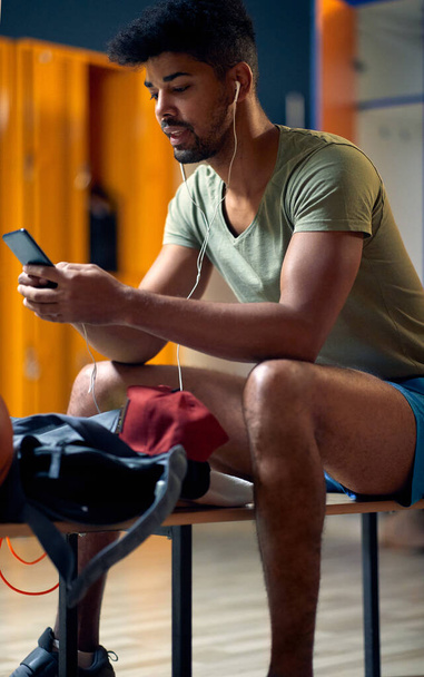 Handsome young sporty man with earphones and smartphone sitting in gym locker room, getting ready for workout session. Health, lifestyle, wellness concept. - Φωτογραφία, εικόνα