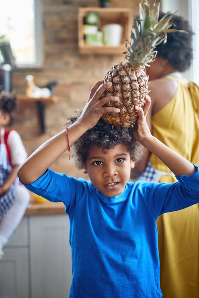 afro-amerykański chłopiec obejmuje jego zabawny duch, gdy stoi w kuchni z niespodziewanym dodatkiem na głowie ananasa - Zdjęcie, obraz