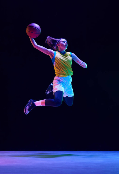 Slam dunk. Basketball player in motion, young girl jumping with ball against black studio background in neon light. Concept of professional sport, action and motion, game, competition, hobby, ad - Valokuva, kuva
