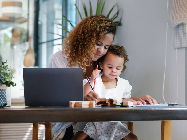 Χαμογελώντας African American μαμά που εργάζονται με φορητό υπολογιστή, ενώ κάθεται ξύλινο τραπέζι στην κουζίνα και παίζει με το κοριτσάκι της. Νεαρή μαύρη μητέρα κάνει freelance το γραφείο με μικρό παιδί στο σπίτι. - Φωτογραφία, εικόνα