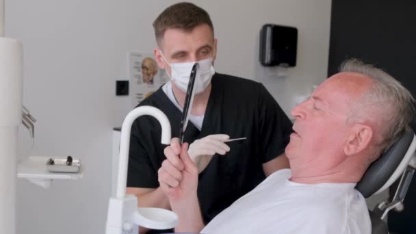 Um pensionista de cabelos grisalhos sentado em uma cadeira de dentista fala sobre seus problemas dentários. Homem dentista no consultório dentário a discutir uma foto dos dentes. Saúde, medicina e odontologia - Filmagem, Vídeo