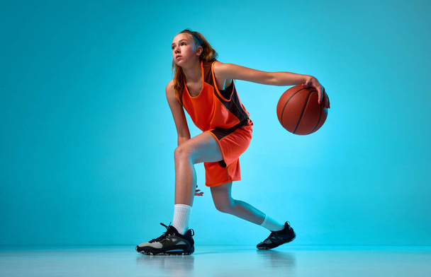 Young athletic girl, basketball player in motion, running with ball against blue studio background in neon light. Concept of professional sport, action and motion, game, competition, hobby, ad - Zdjęcie, obraz