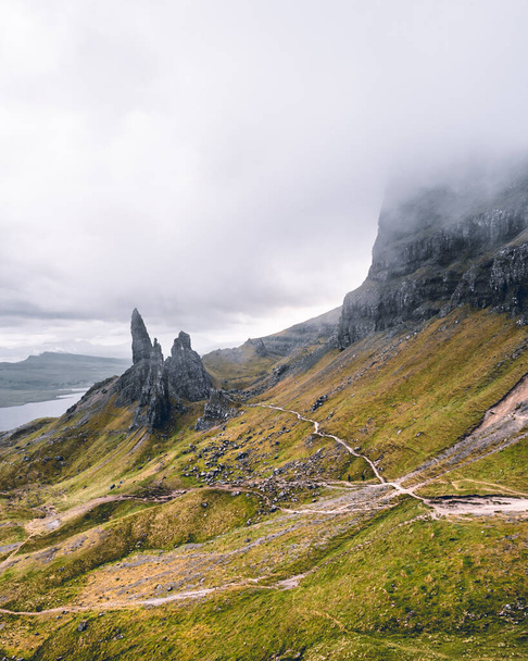 A breathtaking panoramic view reveals the iconic Old Man of Storr in all its glory. Jutting out from the majestic landscape, this ancient rock formation captivates with its towering presence. Surrounded by rolling hills and lush greenery, it stands a - Foto, afbeelding