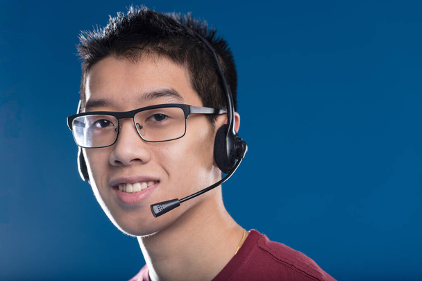 Un joven adulto asiático sonriente usa un jugador típico o auriculares de centro de llamadas y sonríe de manera útil. Los jugadores se comunican con su equipo - Foto, Imagen