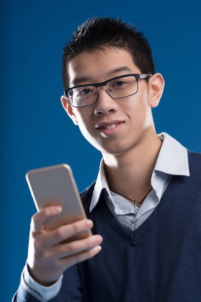 Un hombre asiático sonriente con un smartphone. El uso de gafas, una camisa y un suéter, tiene el pelo corto. Utiliza móviles para aplicaciones, Internet, mensajes, noticias e inteligencias artificiales - Foto, imagen