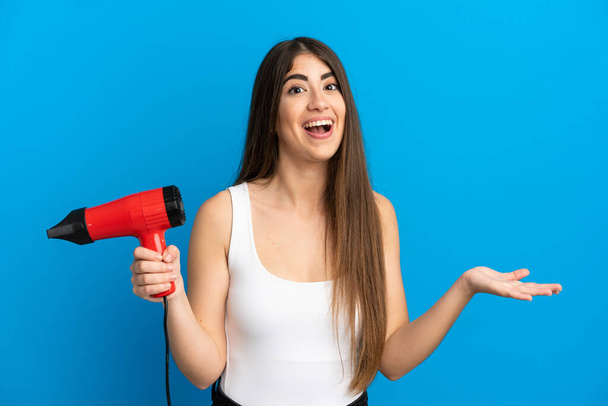 Young caucasian woman holding a hairdryer isolated on blue background with shocked facial expression - Photo, Image