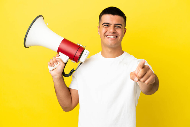 Young handsome man over isolated yellow background holding a megaphone and smiling while pointing to the front - Foto, imagen
