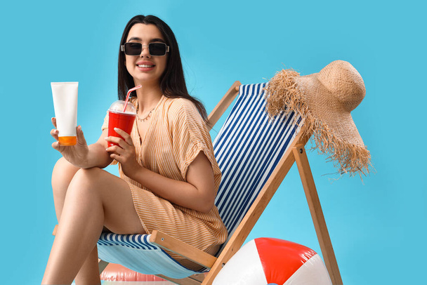 Young woman with sunscreen cream and soda in deck chair on blue background - Foto, Imagem