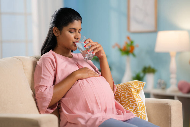 Indische Schwangere trinkt Wasser, während sie zu Hause auf dem Sofa sitzt - Konzept für Hydratation, gesunden Schwangerschaftsstil und Wohlbefinden. - Foto, Bild