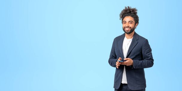 Portrait of cheerful young Middle Eastern businessman in elegant suit holding smartphone standing over blue background. Concept of business and communication. Copy space - Photo, image