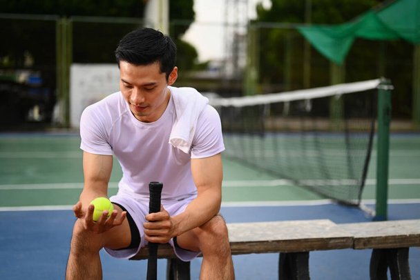 Tired sportsman holding ball racket sitting on the bench at tennis court. Activity, sport and competition concept. - Photo, Image