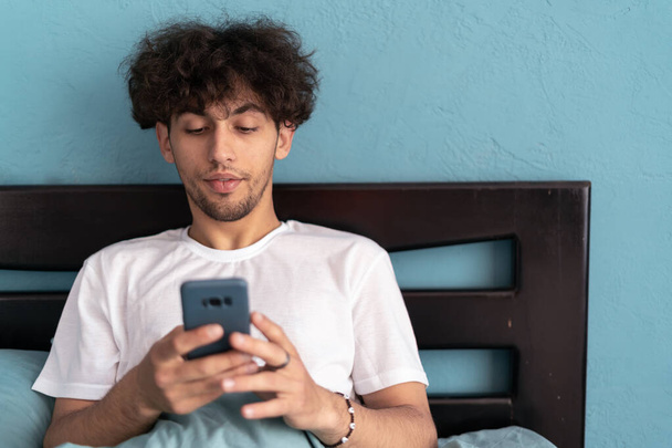 Красивый мужчина, кровать и телефон в домашней спальне для социальных сетей, просмотр интернета по утрам. Технологии, отдых и общение дома. Копирование пространства - Фото, изображение