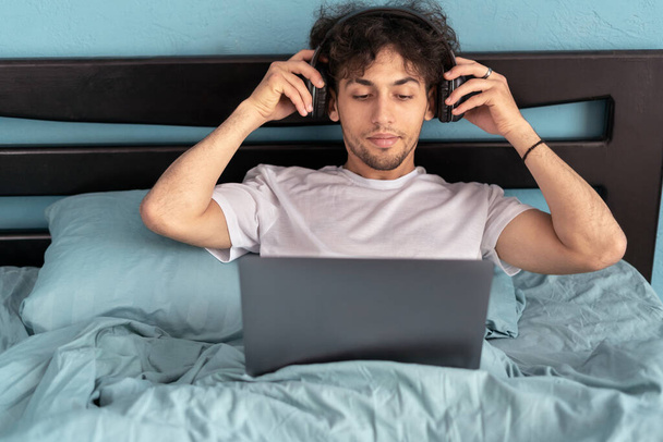 Egy férfi, aki laptopot visel, fülhallgatót visel, filmet néz online, otthon ül az ágyban, a hálószobában. Hétvégi szabadidő és szabadúszó életmód koncepció - Fotó, kép