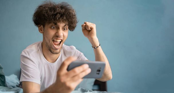 Portret szczęśliwego młodzieńca w białej koszulce patrzącego na smartfona i świętującego zwycięstwo w domowym wnętrzu. Przestrzeń kopiowania - Zdjęcie, obraz