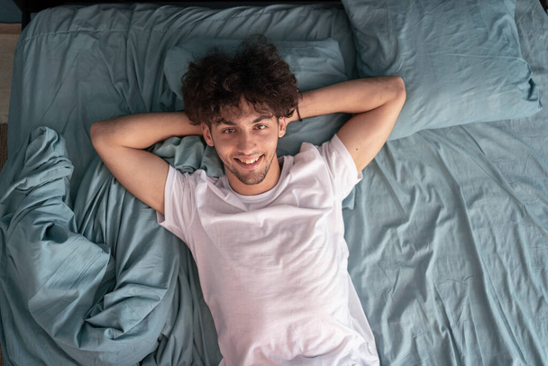 Ein junger lächelnder Mann trägt ein weißes T-Shirt, die Hände unter dem Kopf zusammengefaltet im Bett liegend, blickt in die Kamera, verbringt entspannte Zeit im Schlafzimmer, wacht träumerisch auf. Kopierraum - Foto, Bild