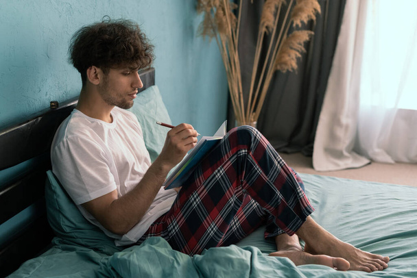 Bello giovanotto prendere appunti nel quaderno mentre sdraiato sul letto scrivere idee e fare pianificazione. Copia spazio - Foto, immagini