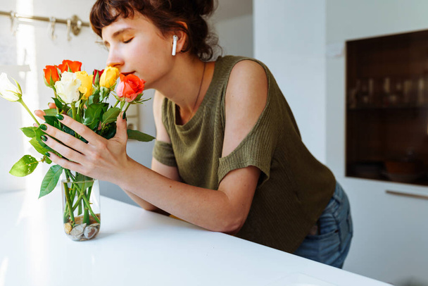 Attractive slender teenager girl, caring for cut flowers roses, bouquet, replacing water in vase, standing in kitchen at home. Pruning stem, arranging bouquet - Photo, Image