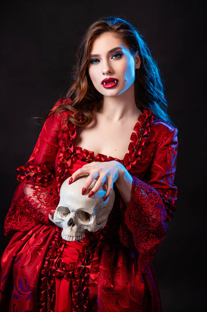 Portrait of a young, attractive vampire woman in a red dress in the rococo era, posing with a skull in her hands isolated on a dark background with blue backlight. - Zdjęcie, obraz