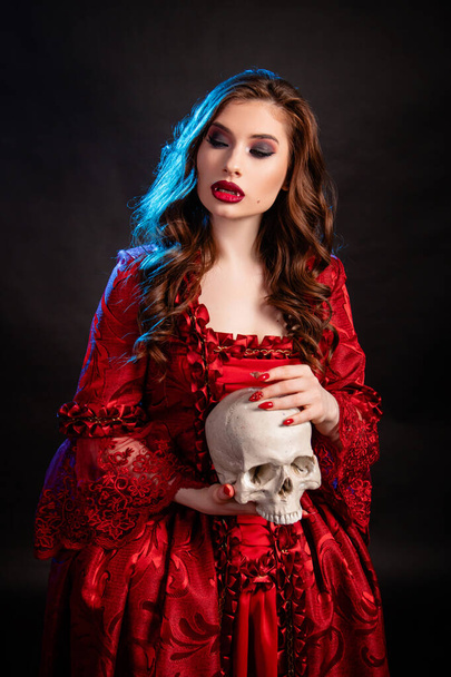 Portrait of a young, attractive vampire woman in a red dress in the rococo era, posing with a skull in her hands isolated on a dark background with blue backlight. - Fotó, kép