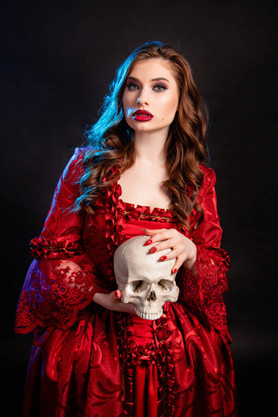 Portrait of a young, attractive vampire woman in a red dress in the rococo era, posing with a skull in her hands isolated on a dark background with blue backlight. - Foto, Imagen