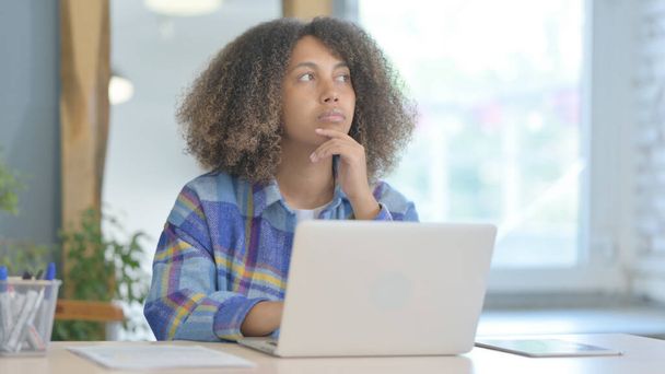 Νεαρή Αφρικανή γυναίκα σκέφτεται και εργάζεται σε φορητό υπολογιστή - Φωτογραφία, εικόνα