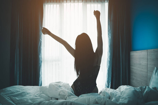 Mladá Asiatka se probudí se šťastným relaxačním pohledem na okno a natahuje paže na posteli v ložnici interiéru. žena vstávat brzy ráno v útulné ložnici. - Fotografie, Obrázek