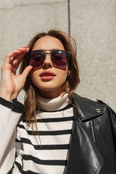 Солнечный городской женский портрет красивой модницы в стильной одежде с свитером и кожаной курткой надевает модные солнцезащитные очки - Фото, изображение