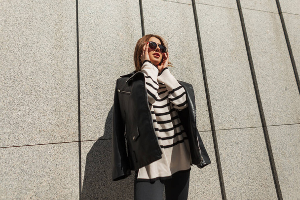 Stylish beautiful young woman model in fashionable clothes with a black leather jacket and sweater puts on sunglasses near the wall in the city on a sunny day, Urban fashion outfit - Foto, imagen