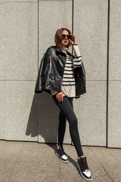 Fashionable beautiful young girl model in stylish clothes with a trendy leather jacket, striped sweater, leggings and sneakers puts on sunglasses on a sunny day near the wall in the city - Photo, image