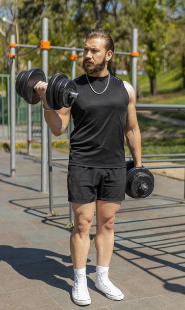 Athletic lebanese man in sportswear doing arms weight lifting dumbbells exercises, pumping up arm bicep muscles. Young guy on playground. Sports, health, fitness routine, workout. Strength motivation - Photo, Image