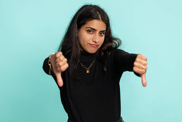 Dislike. Upset unhappy indian woman in sweater showing thumbs down sign gesture, expressing discontent, disapproval, dissatisfied, dislike. Pretty young girl. Indoor studio shot on blue background - Photo, Image