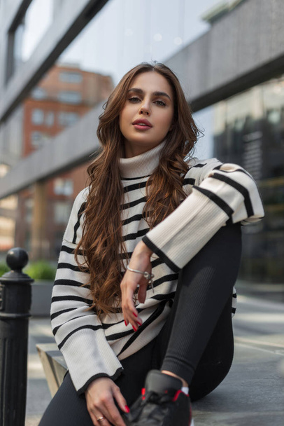 Fashion beauty young stylish woman model with fashionable striped sweater sits and poses in the city - Foto, Imagen