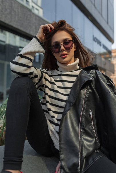 Fashionable young beautiful cool woman with trendy sunglasses in fashion outfit with sweater and black leather jacket sits and poses on the street - Foto, imagen