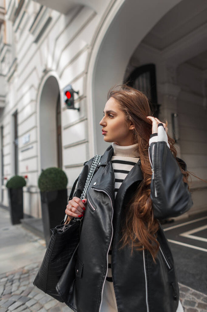 Elegante hermosa mujer joven modelo de moda en ropa de rock casual de moda con un suéter a rayas, chaqueta de cuero negro y paseos bolsa en la ciudad - Foto, Imagen