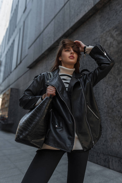 Modieus jong mooi meisje in fashion urban rock outfit met zwarte leren jas en trui met tas wandelingen in de stad in de buurt van een zwart modern gebouw - Foto, afbeelding