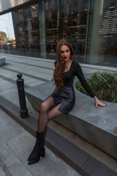 Модная сексуальная женщина с красными губами и стройными ногами в модной элегантной одежде с юбкой, колготками и каблуками сидит в городе - Фото, изображение