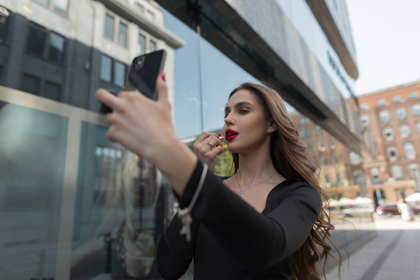 Prachtige mooie jonge mode chique meisje model schildert haar lippen met rode lippenstift en kijkt in een smartphone in de stad - Foto, afbeelding