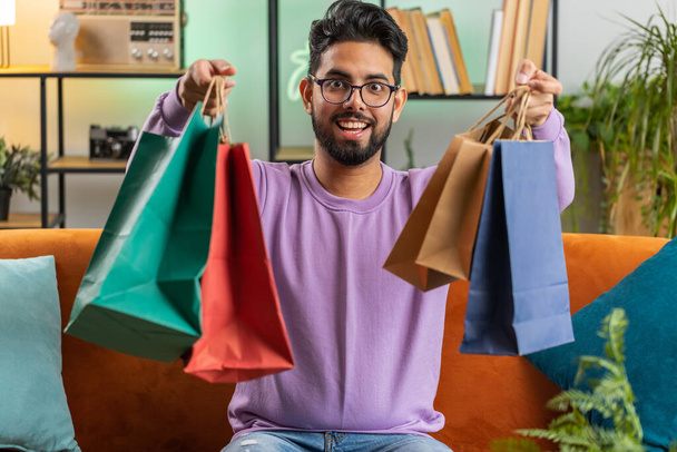 Nuori intialainen mies onnellinen shopaholic kuluttaja palasi kotiin ostosten jälkeen myynti laukkuja. Hindu kaveri tyytyväinen sai paketteja ostaa online järjestyksessä kotona huoneisto huone sohvalla. Pystysuora - Valokuva, kuva