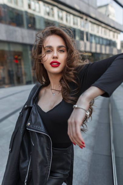 Happy beautiful fashionable chic girl with red lips in a stylish elegant outfit with a leather jacket poses in the city on a windy day - Φωτογραφία, εικόνα