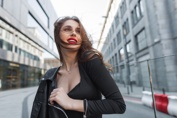Happy beautiful fashion lady with red lips in trendy stylish black clothes with leather jacket walking in the city on a windy day - Foto, immagini